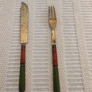 May include: A vintage knife and fork set. The knife has a gold-coloured blade and a handle with red and green sections. The fork is gold-coloured with three tines and a matching handle. The set is likely antique and shows signs of wear.