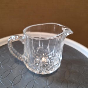 May include: A clear glass creamer with a handle and a spout. The creamer has a cut-glass design, with a textured pattern on the body and handle. The creamer is designed to hold liquids and is suitable for serving.
