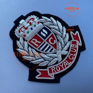 Op de afbeelding: Een zwart geborduurd embleem met een kroon, een lauwerkrans en een schild met de letters "RC". Een rode banner met "ROYAL CLUB" is eronder genaaid. De patch is gedetailleerd met zilveren draad.