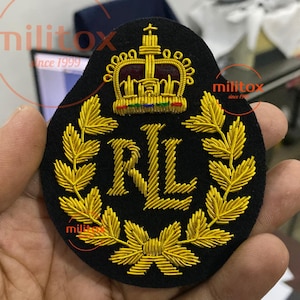 Pode incluir: Um patch preto com bordado dourado apresentando uma coroa, uma coroa de louros e as letras "RL". O patch tem formato circular e é provavelmente um emblema militar ou formal.