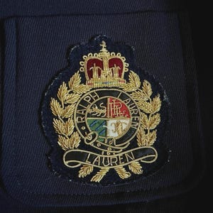 Peut inclure: Un vêtement en tissu bleu marine foncé orné d'un écusson brodé. L'écusson présente une couronne dorée au-dessus d'un emblème circulaire avec le texte RALPH LAUREN, entourant un bouclier divisé en quatre quadrants aux détails dorés, rouges et verts.
