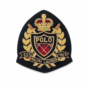 Op de afbeelding: Een zwart, schildvormig geborduurd embleem met een gouden kroon boven een wapenschild met de tekst POLO en een rood ruitpatroon. Het ontwerp bevat gouden lauriertakken, gekruiste zwaarden en een banner met de tekst EST. 1967 RALPH LAUREN.