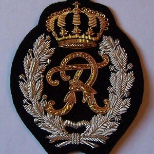May include: A black embroidered patch with a gold crown and a gold letter "R" intertwined with a gold design. Silver-colored laurel leaves surround the letter and crown. The patch has a black background.