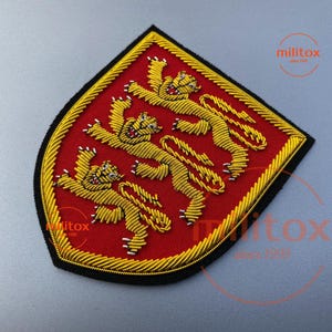 May include: Embroidered patch featuring three gold lions rampant on a red background, framed by a gold border and a black edge. The patch has a shield shape and is likely a military or heraldic emblem.