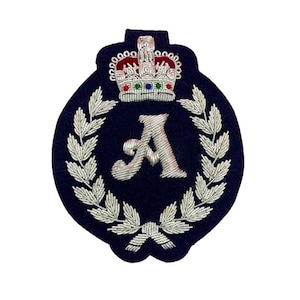 May include: A navy blue embroidered patch featuring a silver-colored letter "A" within a laurel wreath. A detailed silver crown with red, green, and blue accents sits above the letter. The patch has a dark blue oval shape.