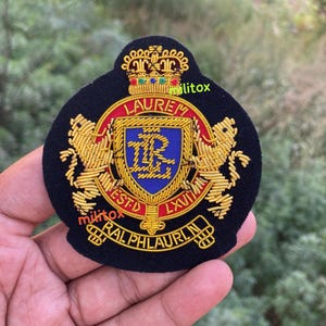 May include: A black embroidered patch with a gold crown, the word "LAUREN", and a crest with the letters "R" and "L". The patch features gold lions and the words "RALPH LAUREN" and "ESTD LXVII".