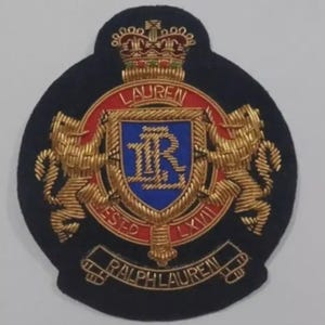 Op de afbeelding: Geborduurde patch met een marineblauwe achtergrond. De patch heeft een centraal schild met de gouden letters "LR", omgeven door een rode ring met het woord "LAUREN" bovenaan. Twee gouden leeuwen flankeren het schild. De woorden "ESTD LXVII" en "RALPH LAUREN" zijn ook geborduurd.
