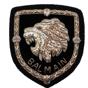 Puede incluir: Parche negro en forma de escudo con un diseño de león rugiente. El león está bordado con hilos plateados y dorados. La palabra "BALMAIN" está bordada en oro en la parte inferior. El borde también es dorado y plateado.