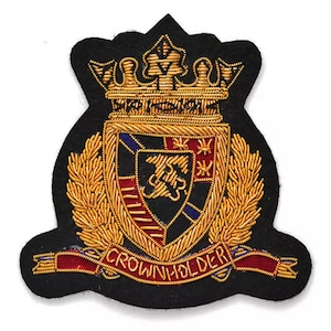 Op de afbeelding: Geborduurde patch met een heraldisch wapen. Het heeft een gouden kroon, laurierbladeren en een schild met rode, blauwe en gouden details. De tekst "CROWN HOLDER" staat op een rode banner. Zwarte achtergrond.