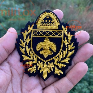 May include: A navy blue embroidered patch with gold thread detailing. The patch features a shield design with a fleur-de-lis and a crown, surrounded by laurel leaves. The text "militox since 1999" is visible.