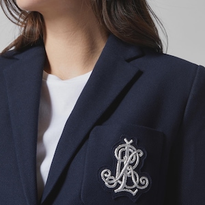 May include: A navy blue blazer with a textured fabric finish, featuring a white embroidered monogram patch on the chest pocket. The blazer has a notched lapel and a silver metallic button visible on the front, worn over a plain white shirt.