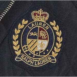 以下が含まれることがあります： ラルフローレンのエンブレムが付いたネイビーブルーのパッチ。円形のエンブレムは、金色の月桂樹のリースが付いています。ネイビーブルーの中心部には、王冠、「LAUREN」の文字、さまざまなシンボルが描かれたシールドデザインが含まれています。