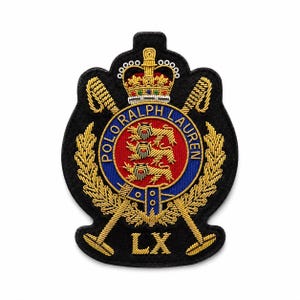 Könnte beinhalten: Ein schwarzer gestickter Aufnäher mit einer goldenen Krone über einem roten kreisförmigen Wappen mit drei Löwen. Das Wappen ist von einem blauen Rand mit dem Text POLO RALPH LAUREN in Gold umgeben, flankiert von zwei gekreuzten goldenen Poloschlägern und den Buchstaben LX.