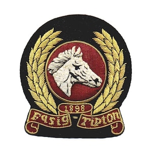 Puede incluir: Parche bordado con fondo negro, con una cabeza de caballo blanca dentro de un círculo rojo. Hojas de laurel doradas rodean el círculo. El parche incluye el texto "1898" y "Paris Ripton" en dorado.