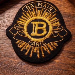 Könnte beinhalten: Schwarz-goldener, gestickter Aufnäher mit dem Wort "BALMAIN" oben und "PARIS" unten. In der Mitte befindet sich ein goldenes "B" in einem goldenen Kreis. Der Aufnäher liegt auf einer Holzoberfläche.