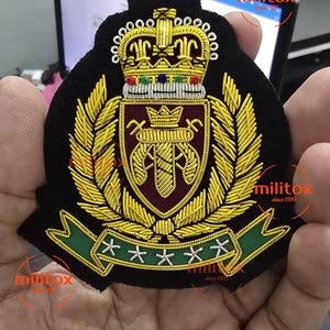 Op de afbeelding: Een zwart geborduurd embleem met een gouden kroon, laurierbladeren en een wapen met een bordeauxrode achtergrond. Het embleem heeft een groene banner met witte sterren. De tekst "militox since 1999" is zichtbaar.