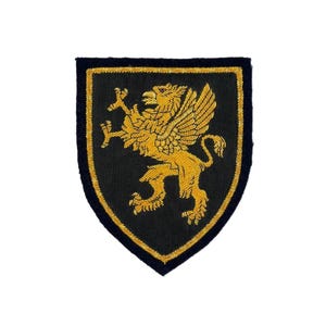 May include: A black and gold embroidered patch featuring a heraldic griffin design. The griffin is gold with outstretched wings, set against a black shield shape with a gold border. The patch is likely for apparel or accessories.