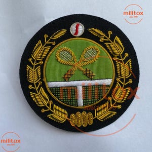 Può includere: Toppa ricamata rotonda con tema tennis. La toppa ha una base nera con foglie d'alloro dorate. Il centro presenta racchette da tennis incrociate, una rete e uno sfondo verde. Il testo "militox since 1999" è visibile.