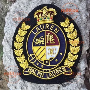 Op de afbeelding: Een zwarte geborduurde patch met een gouden kroon aan de bovenkant en gouden lauriertakken aan de zijkanten. Het centrale ronde embleem toont de tekst LAUREN en RALPH LAUREN met een schild verdeeld in vier kwadranten met een leeuw en letters.