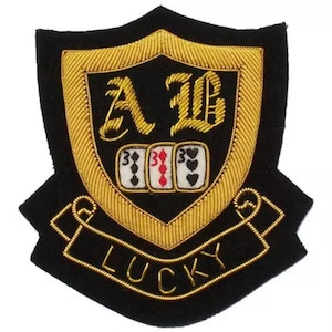 May include: Black and gold embroidered patch with a shield design. The patch features the letters "A" and "B", three playing card symbols, and the word "LUCKY". The border and lettering are gold, and the background is black.