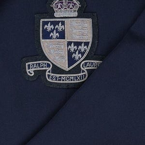Puede incluir: Primer plano de una prenda azul marino con un escudo bordado. El escudo incluye una corona, un escudo con diseños florales y las palabras "RALPH LAUREN" y "EST-MCMLXII" en una pancarta.