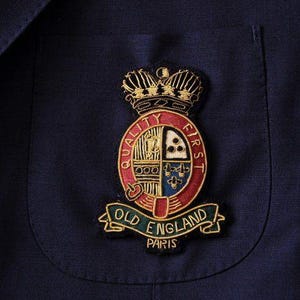 Op de afbeelding: Close-up van een marineblauwe blazer met een zak. De blazer heeft een geborduurd embleem met een kroon, een rood-gouden embleem en de tekst "OLD ENGLAND PARIS". De patch bevindt zich in de linker bovenpocket.