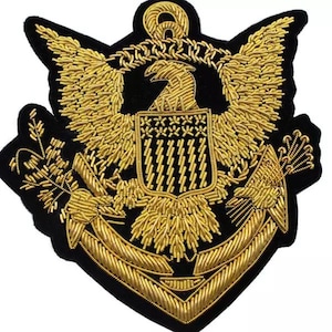 Op de afbeelding: Een goudkleurige geborduurde patch met een adelaar met uitgespreide vleugels, een schild en een anker. De patch staat op een zwarte achtergrond. De adelaar is het centrale punt, met ingewikkelde details in gouddraad. De patch is waarschijnlijk een militair of nautisch embleem.