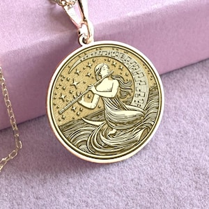 Op de afbeelding: Goudkleurige hangerketting met een gedetailleerde gravure van een vrouw die fluit speelt, omringd door sterren en muzieknoten. De hanger hangt aan een delicate ketting. Het ontwerp is geplaatst tegen een zachtroze achtergrond.