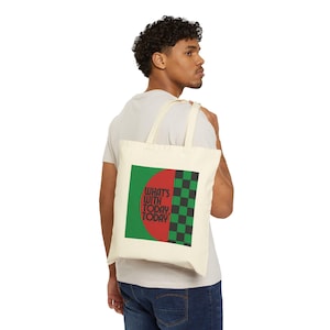 May include: A cream-coloured tote bag with a graphic design. The design features the text "WHAT'S WITH TODAY, TODAY" in red and white, set against a green square and a black and green chequered pattern. The bag is carried over the shoulder.