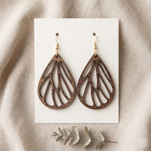 May include: A pair of teardrop-shaped earrings crafted from dark brown wood, featuring an intricate cutout pattern resembling insect wings. The earrings are suspended from gold-toned metal fishhook wires and are displayed on a white card.