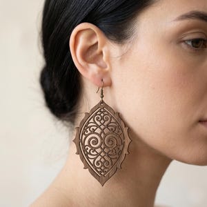 May include: A large, teardrop-shaped wooden earring featuring an intricate, laser-cut filigree pattern with symmetrical scrollwork. The earring is crafted from dark brown wood and hangs from a simple, thin bronze-toned metal hook wire.