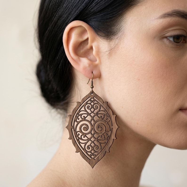 May include: A large, teardrop-shaped wooden earring featuring an intricate, laser-cut filigree pattern with symmetrical scrollwork. The earring is crafted from dark brown wood and hangs from a simple, thin bronze-toned metal hook wire.