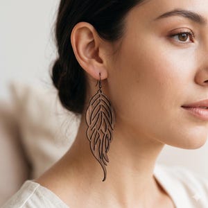 May include: A dark brown wooden dangle earring featuring a laser-cut feather or leaf design with intricate internal cutouts. The earring hangs from a thin, dark metal hook, showcasing a natural wood grain texture and a long, slender, curved shape.