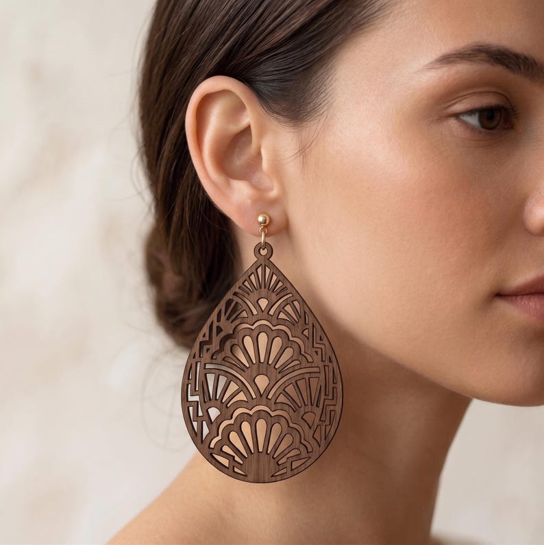 May include: A teardrop-shaped wooden earring featuring a dark brown finish and an intricate laser-cut pattern of repeating fan-like arches. The earring is attached to a small gold-toned metal stud post that secures it to the earlobe.