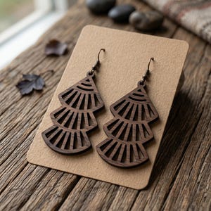 May include: A pair of dark brown wooden earrings featuring a tiered, fan-like geometric cutout design. Each earring is suspended from a dark metal hook and displays a repeating pattern of radiating lines within three stacked, triangular sections.