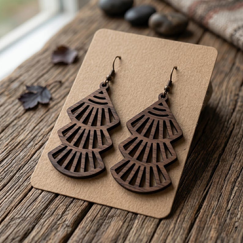 May include: A pair of dark brown wooden earrings featuring a tiered, fan-like geometric cutout design. Each earring is suspended from a dark metal hook and displays a repeating pattern of radiating lines within three stacked, triangular sections.