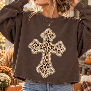 May include: A dark brown long-sleeve sweatshirt featuring a large central cross graphic with a leopard print pattern. The cross is outlined with a border of small white faux pearls and has a distressed, frayed fabric texture throughout.