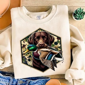 Op de afbeelding: Crèmekleurige sweatshirt met een bruine Labrador Retriever die een wilde eend in zijn bek houdt. De hond en de eend bevinden zich in een groene en bruine zeshoek met een camouflagepatroon. Ronde hals.