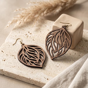 May include: A pair of dark brown wooden drop earrings featuring a laser-cut leaf or teardrop shape with intricate, flowing internal cutouts. Each earring is attached to a gold-toned metal hook for hanging, showcasing a natural wood grain texture.
