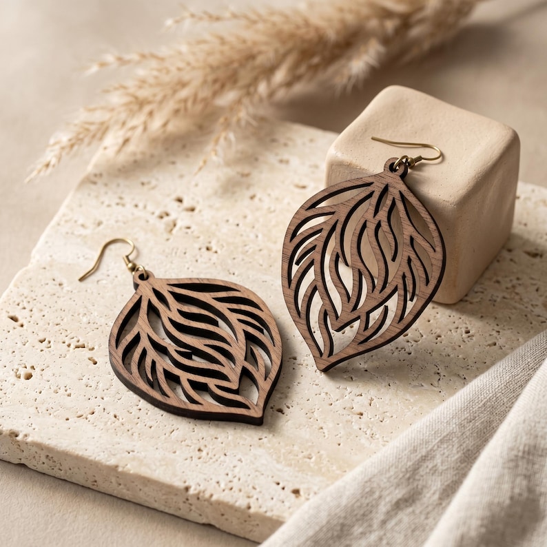 May include: A pair of dark brown wooden drop earrings featuring a laser-cut leaf or teardrop shape with intricate, flowing internal cutouts. Each earring is attached to a gold-toned metal hook for hanging, showcasing a natural wood grain texture.