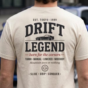 May include: A cream-colored short-sleeve t-shirt featuring black and red text on the back that reads "EST. TOKYO 1989 DRIFT LEGEND born for the corners TURBO MANUAL LOWERED WIDEBODY Mountain pass or nothing SLIDE GRIP CONQUER" with a car graphic.