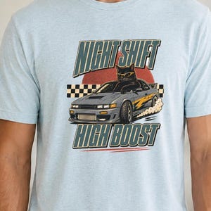 May include: A heather prism ice blue short-sleeve t-shirt featuring a graphic print of a black cat wearing sunglasses driving a grey sports car. The text NIGHT SHIFT and HIGH BOOST appears above and below the car with a checkered flag pattern.