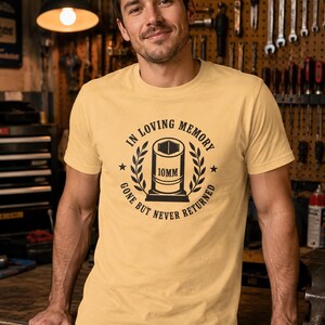 May include: A light yellow short-sleeve t-shirt featuring a black graphic of a 10mm socket surrounded by laurel leaves. The text reads IN LOVING MEMORY and GONE BUT NEVER RETURNED, with a star on each side of the central socket illustration.