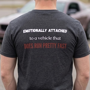 Puede incluir: Una camiseta de manga corta en color gris carbón oscuro con texto en blanco y rojo en la parte posterior que dice: EMOTIONALLY ATTACHED to a vehicle that DOES RUN PRETTY FAST. La prenda tiene cuello redondo y una textura jaspeada.