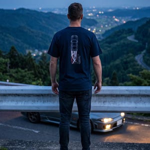 May include: A navy blue t-shirt with a car graphic and Japanese text on the back. The person is standing on a concrete barrier overlooking a mountain road with a car and city lights in the background.