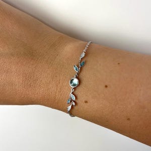 Könnte beinhalten: Ein filigranes Silberarmband mit einem zentralen, hellblauen Edelstein und blattförmigen Akzenten. Das Armband hat einen Kettenverschluss und ist mit kleinen, hellblauen und klaren Edelsteinen verziert, was ein elegantes Design ergibt.
