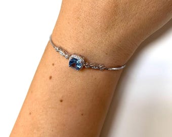 Puede incluir: Una pulsera de plata con una piedra preciosa cuadrada de color azul claro rodeada de pequeñas piedras transparentes. La pulsera tiene una cadena delicada con detalles decorativos. Una joya.