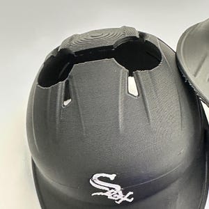 Puede incluir: Casco de b&eacute;isbol negro con el logotipo de los Chicago White Sox en la parte delantera. El casco tiene una superficie texturizada y un dise&ntilde;o &uacute;nico con aberturas en la parte superior. La visera es curva.