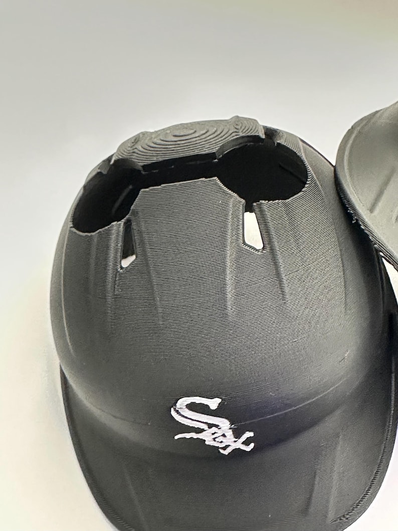 Puede incluir: Casco de b&eacute;isbol negro con el logotipo de los Chicago White Sox en la parte delantera. El casco tiene una superficie texturizada y un dise&ntilde;o &uacute;nico con aberturas en la parte superior. La visera es curva.
