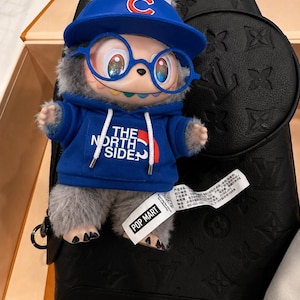 May include: A plush toy figure wearing a blue Chicago Cubs baseball cap and a blue hoodie with "THE NORTH SIDE" printed on it. The figure has blue glasses and is set against a black leather bag.